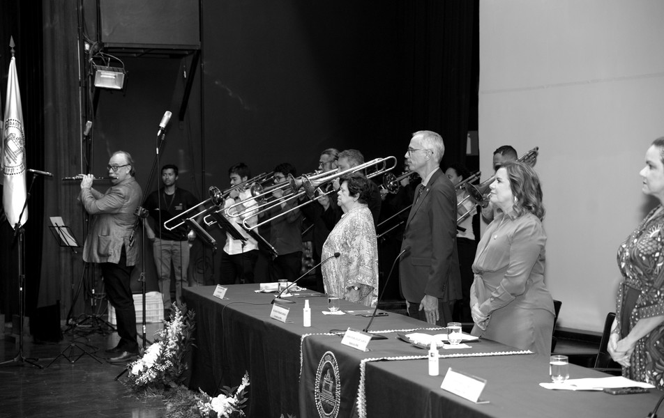 <p>Maurício Freire, pró-reitor de Planejamento e&nbsp;professor da Escola de Música,&nbsp;executa o Hino Nacional acompanhado pelo&nbsp;Coral de Trombones da UFMG.&nbsp;Na ponta da mesa, Ana Lúcia Gazzola, reitora na gestão 2002-2006</p>